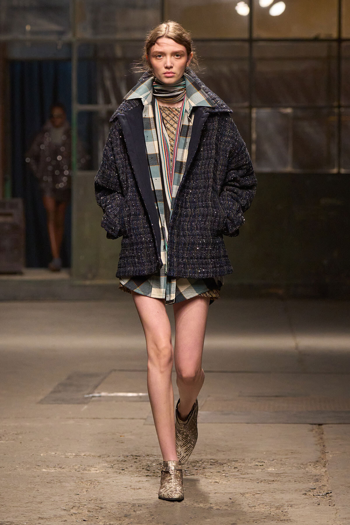 Model walks the runway in a dark, oversized textured navy coat layered over plaid pieces and ankle boots.