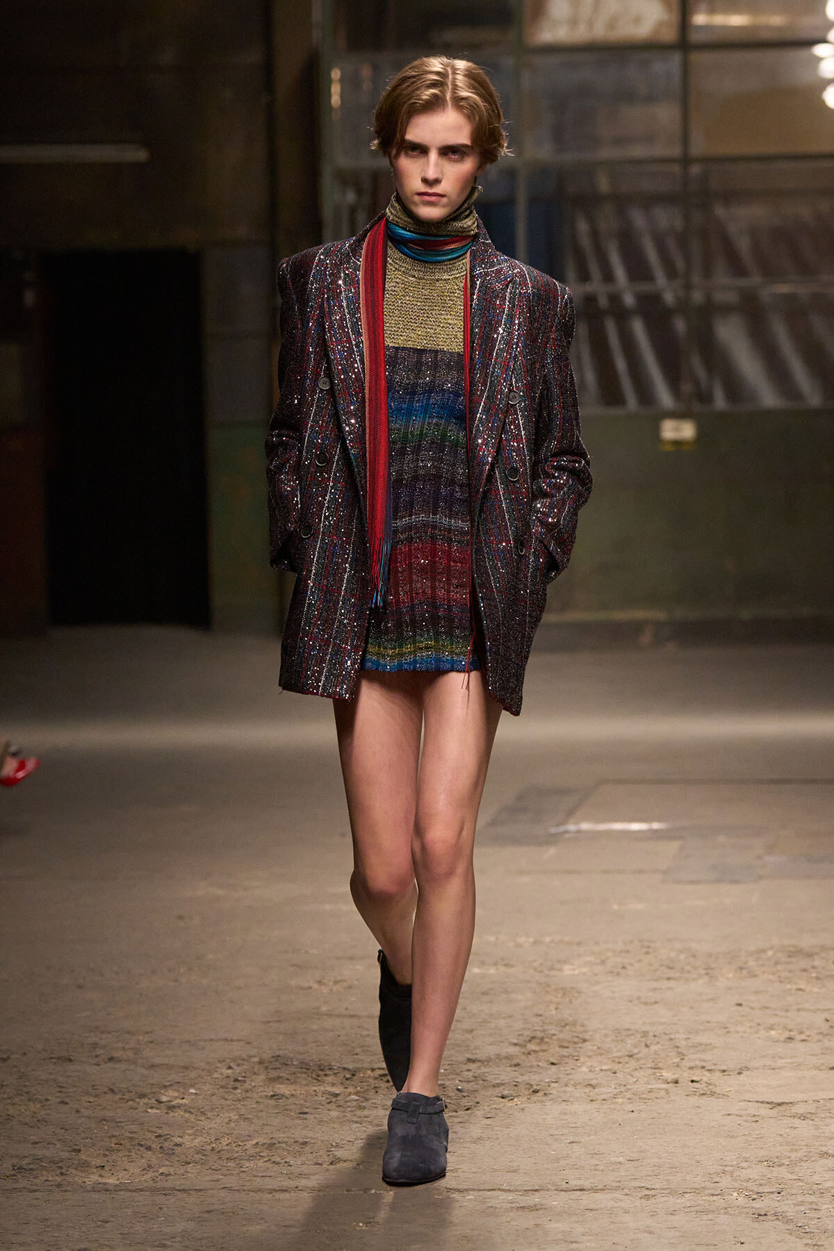Model walking a runway in a metallic, sequined blazer over a multicolor striped knit dress with a red scarf.