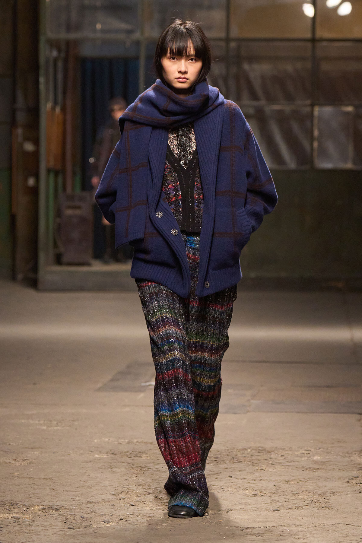 Model walks a runway in an oversized navy plaid cardigan with a matching scarf and multicolor knitted trousers.