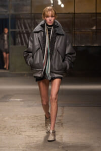Model walking a runway in an oversized gray leather bomber jacket with a striped scarf and neutral boots.