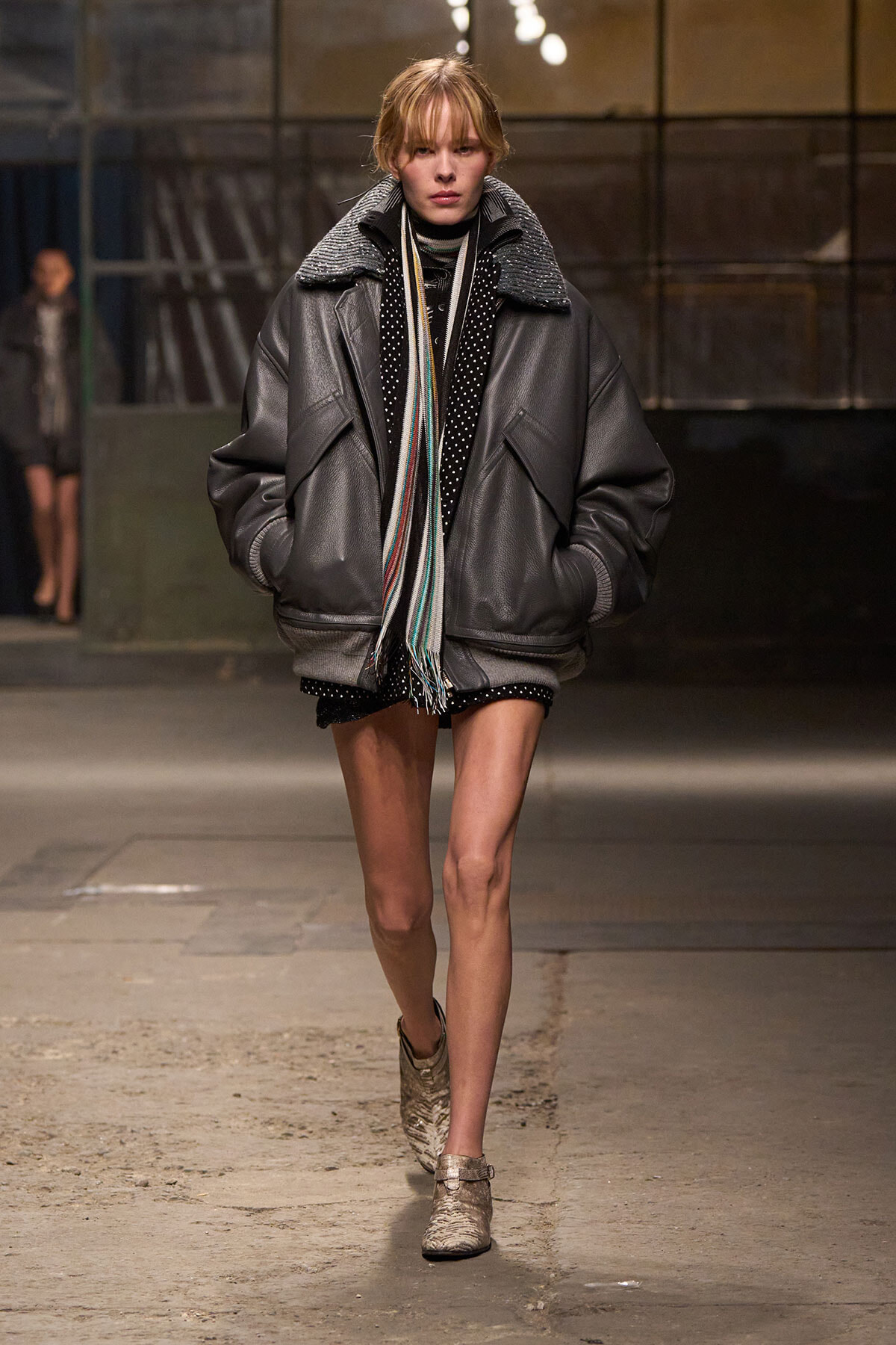 Model walking a runway in an oversized gray leather bomber jacket with a striped scarf and neutral boots.