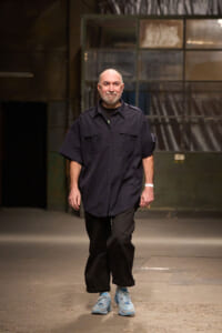 Man walking toward the camera in a dim industrial space, wearing a navy short-sleeve button-up shirt, black pants, and light blue sneakers.