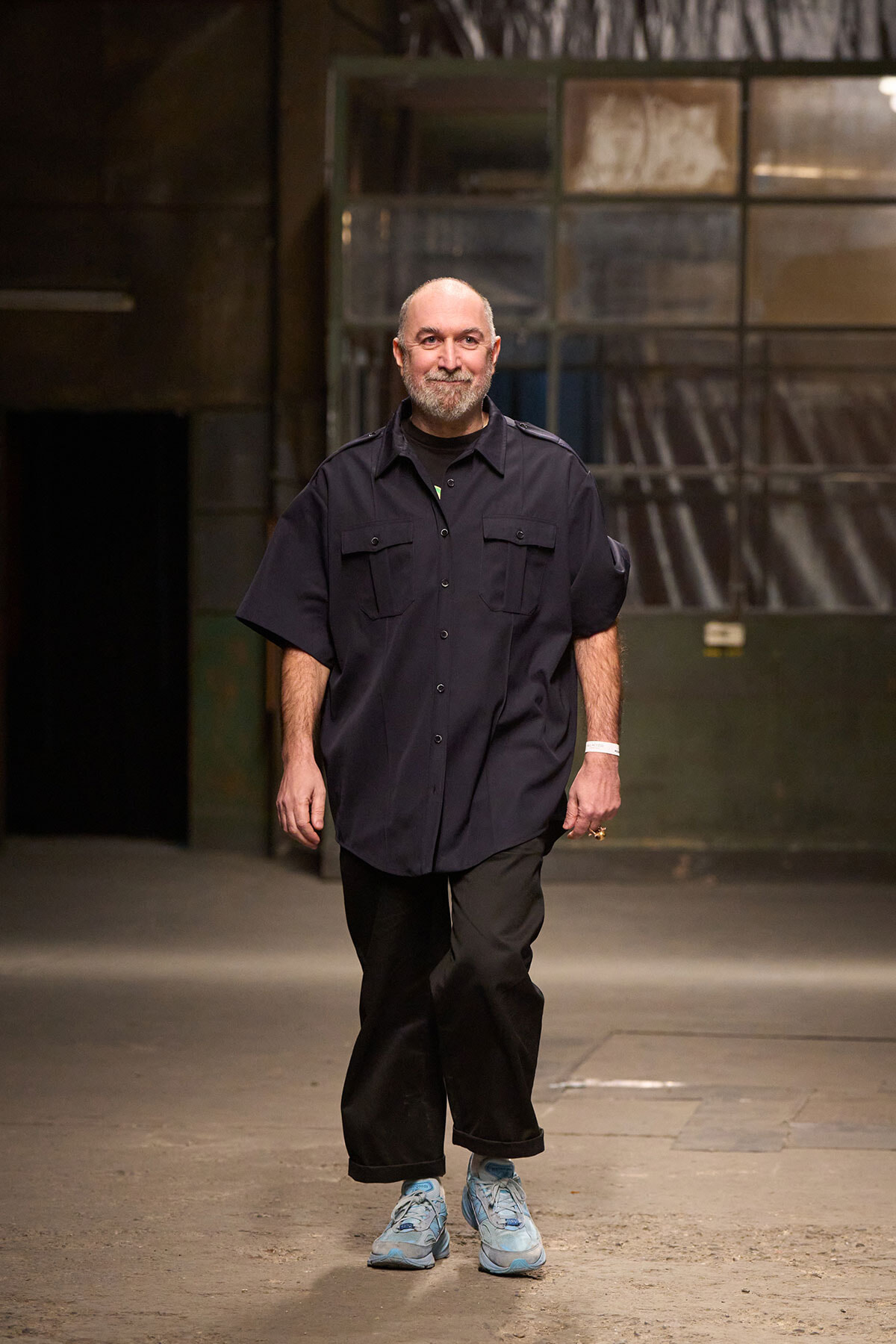 Man walking toward the camera in a dim industrial space, wearing a navy short-sleeve button-up shirt, black pants, and light blue sneakers.