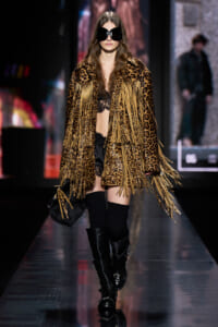 Model on a runway wearing a leopard-print fringe coat, black lingerie, thigh-high socks, and tall black boots with oversized sunglasses.