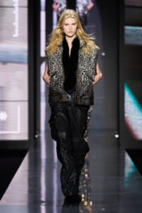 Female runway model walking the catwalk in a studded, silver-embellished vest with black fur trim over a dark outfit and black cargo-style pants.