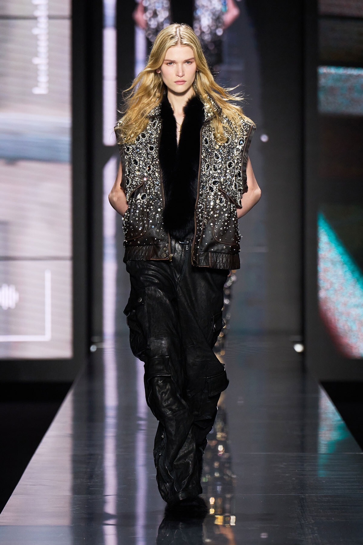 Female runway model walking the catwalk in a studded, silver-embellished vest with black fur trim over a dark outfit and black cargo-style pants.