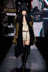 Model on a runway in a black knit beanie, oversized coat with fur panels, a beige lace slip dress, and tall black buckled boots.
