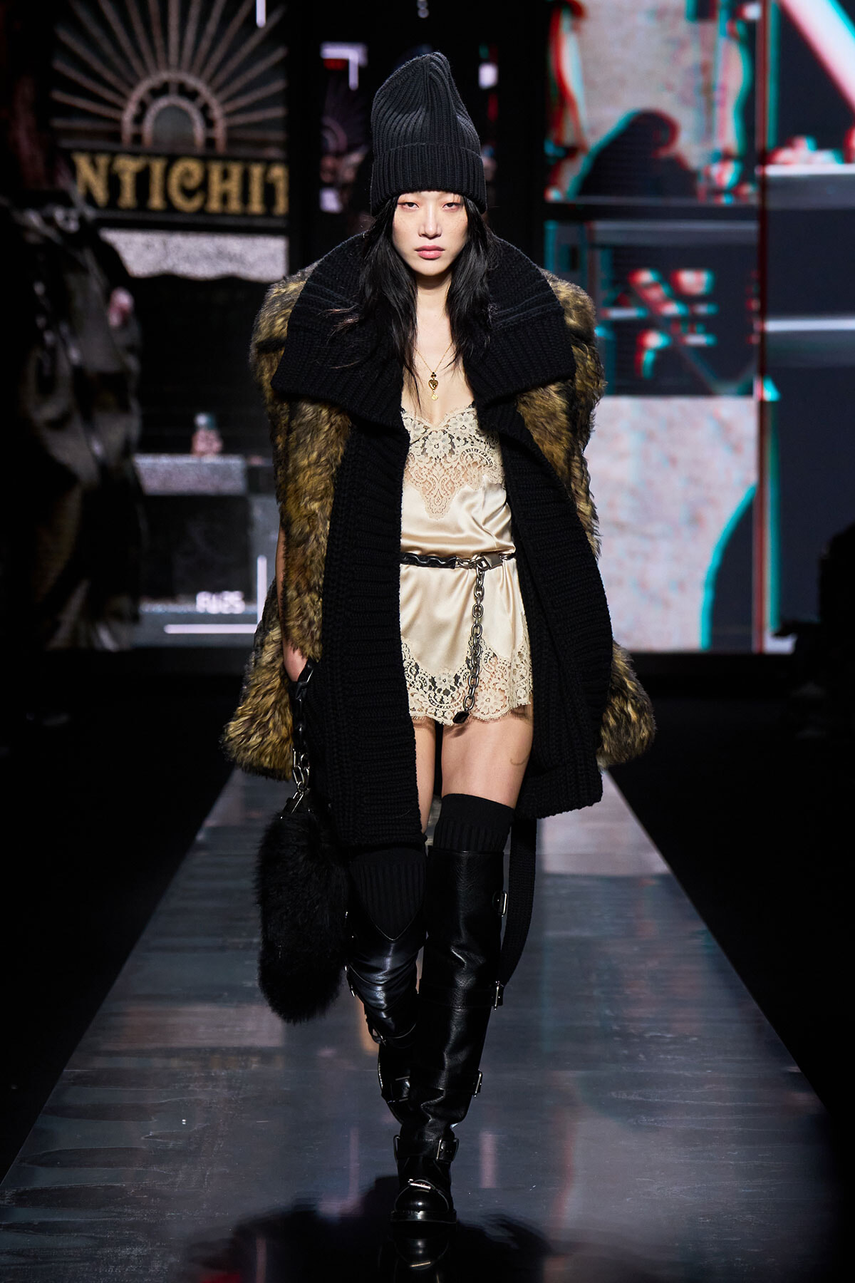 Model on a runway in a black knit beanie, oversized coat with fur panels, a beige lace slip dress, and tall black buckled boots.
