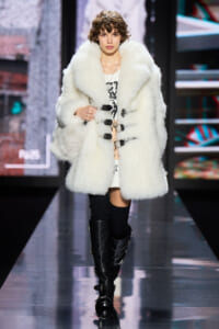 Model walking the runway in a large white fur coat with black buckle closures, paired with a white dress and tall black boots.