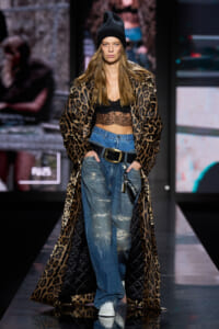 Model walking a runway in a leopard-print long coat over a black lace crop top, distressed jeans with a wide belt, white sneakers, and a black knit beanie.