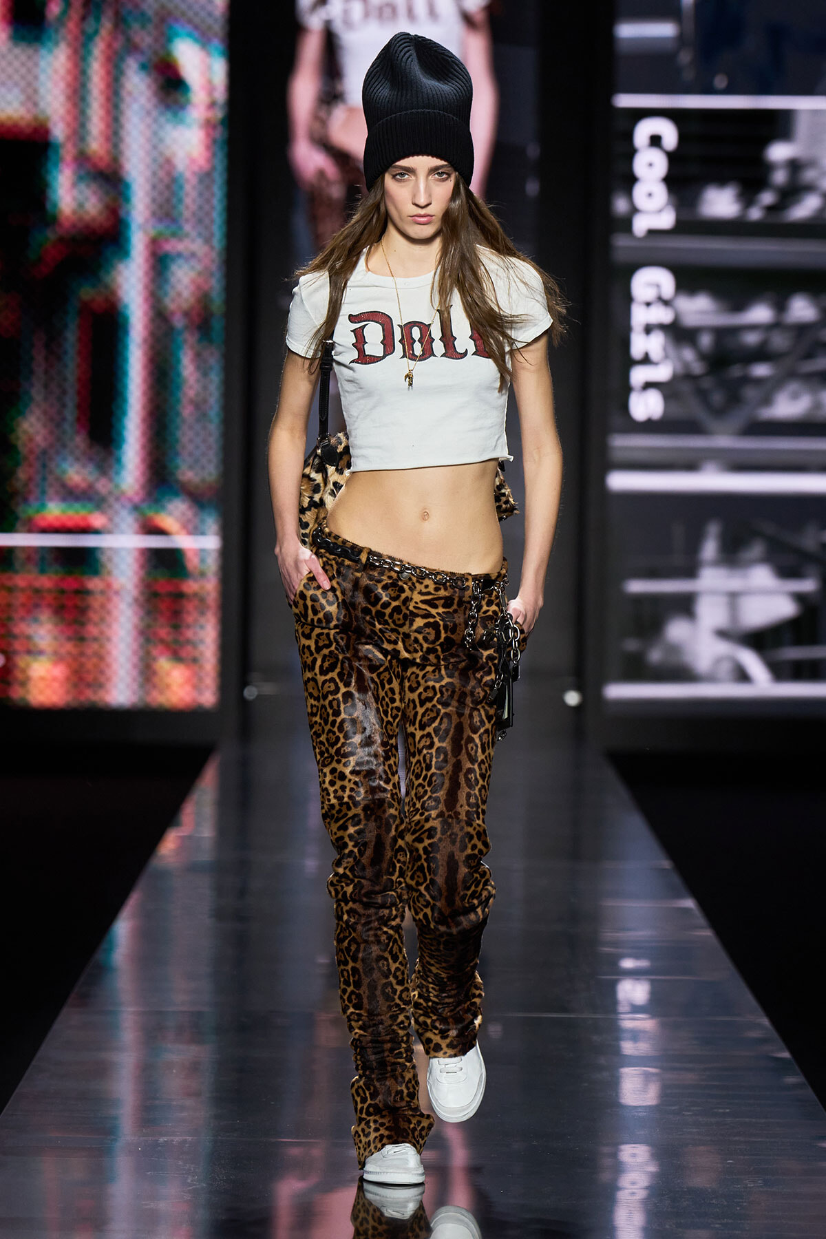 Model walking a fashion show in leopard print pants, cropped white top with 'Doll', black beanie, and white sneakers on a glossy runway.