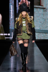 Model walks a fashion runway wearing an olive green tunic with a fur-trim vest, black knit sleeves, lace shorts, and knee-high boots while carrying a brown handbag.