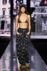 Runway model wearing a black lace bralette, embroidered wide-leg pants, chain belt, and a dark knit beanie with furry boots on a glossy catwalk.