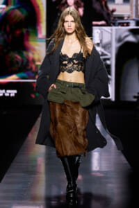 Model walks the runway in a black lace bralette, oversized dark cardigan, olive cargo belt with pockets, brown fur skirt, and tall black boots.