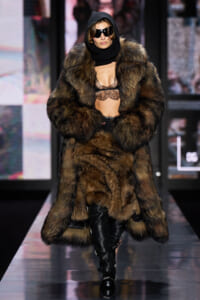 Model on a fashion runway wearing a brown fur coat with hood, black lace bralette, scarf, and oversized sunglasses.
