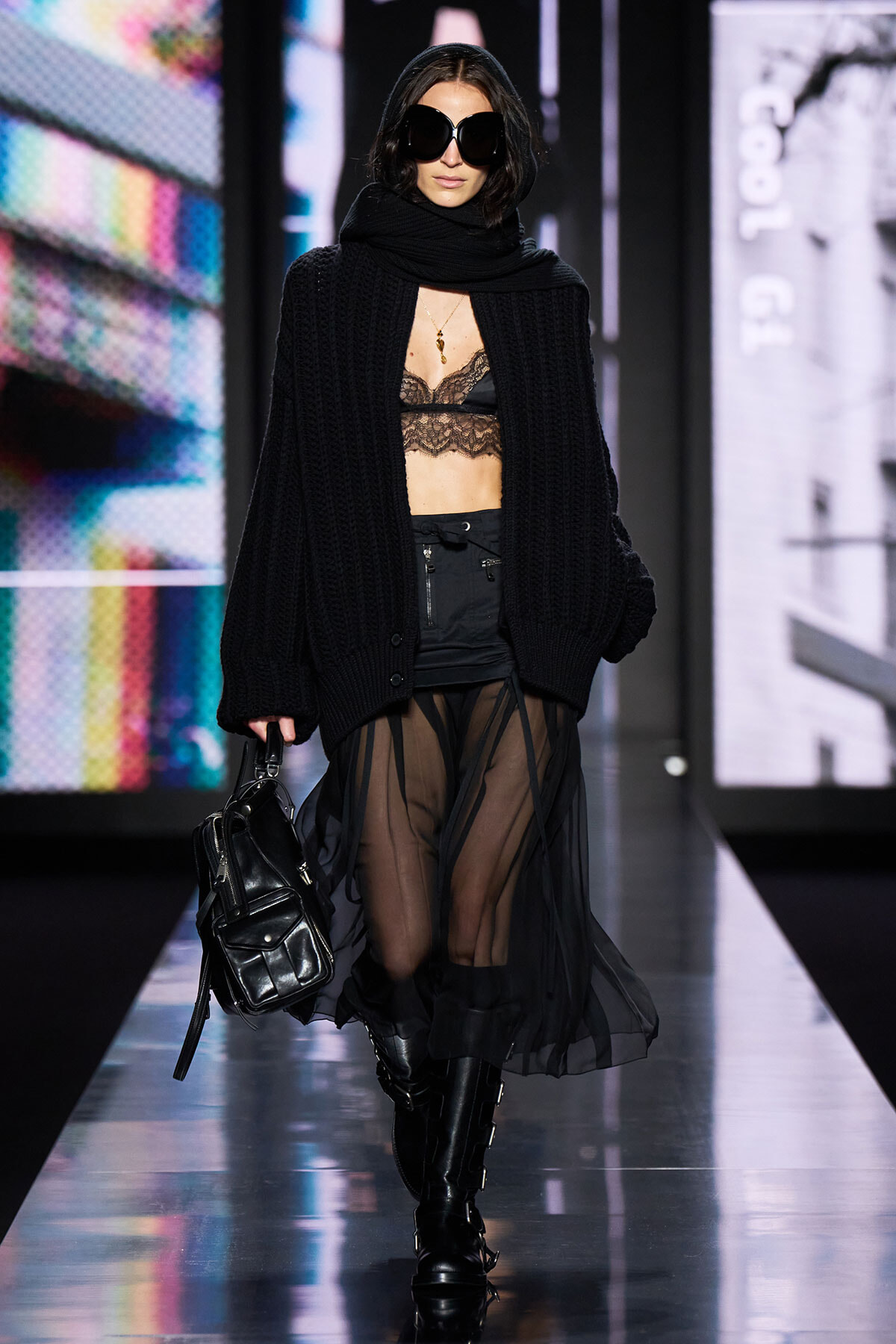 Model on a runway in an all-black look: oversized knit cardigan, lace bralette, sheer black skirt, knee-high boots, large sunglasses, and a black handbag.