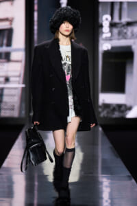 Model walks a fashion runway in an oversized black coat, furry hat, graphic dress, and fur-trimmed boots, carrying a black backpack.