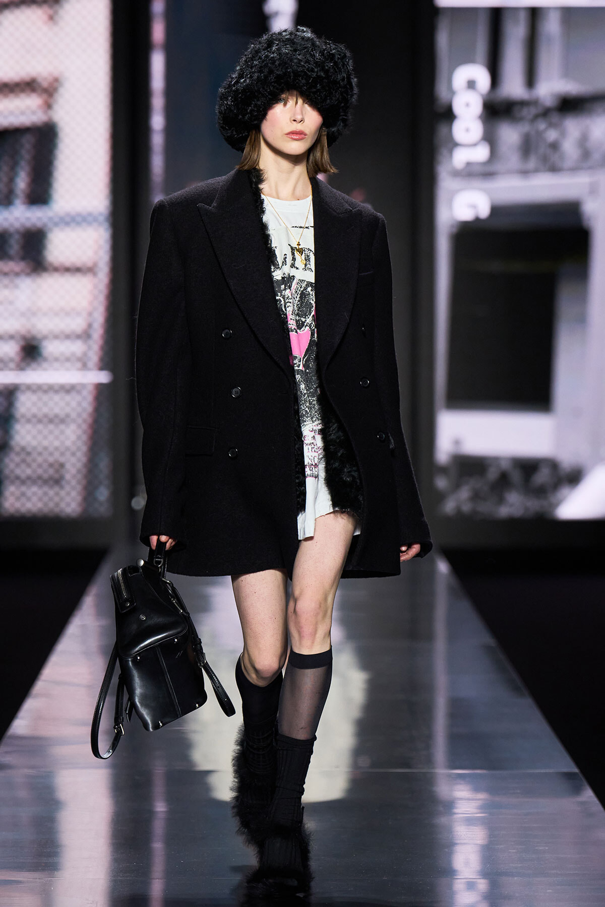 Model walks a fashion runway in an oversized black coat, furry hat, graphic dress, and fur-trimmed boots, carrying a black backpack.