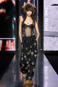 Model walks the runway in a sheer black lace bodice with a fur hat, paired with oversized black pants covered in jewel-like appliqués and furry boots.