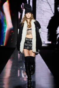 Model walking the runway in a black varsity jacket with white shearling trim, lace bralette, and a sequined mini skirt, paired with tall black boots and a knit beanie.