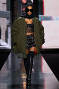 Model walking a fashion runway in an oversized olive bomber jacket, black lace lingerie top, and black satin shorts, paired with knee-high buckled boots and a black headscarf and oversized sunglasses.