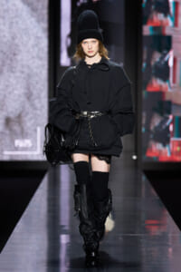 Model walking a runway in an oversized black hoodie, beanie, knee-high socks, and black combat boots, carrying a black bag with a chain belt.