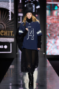 Runway model in a navy oversized jersey with 84, black knit layers, a sheer black lace skirt, tall black boots, beanie, and a black fur coat over the shoulder.