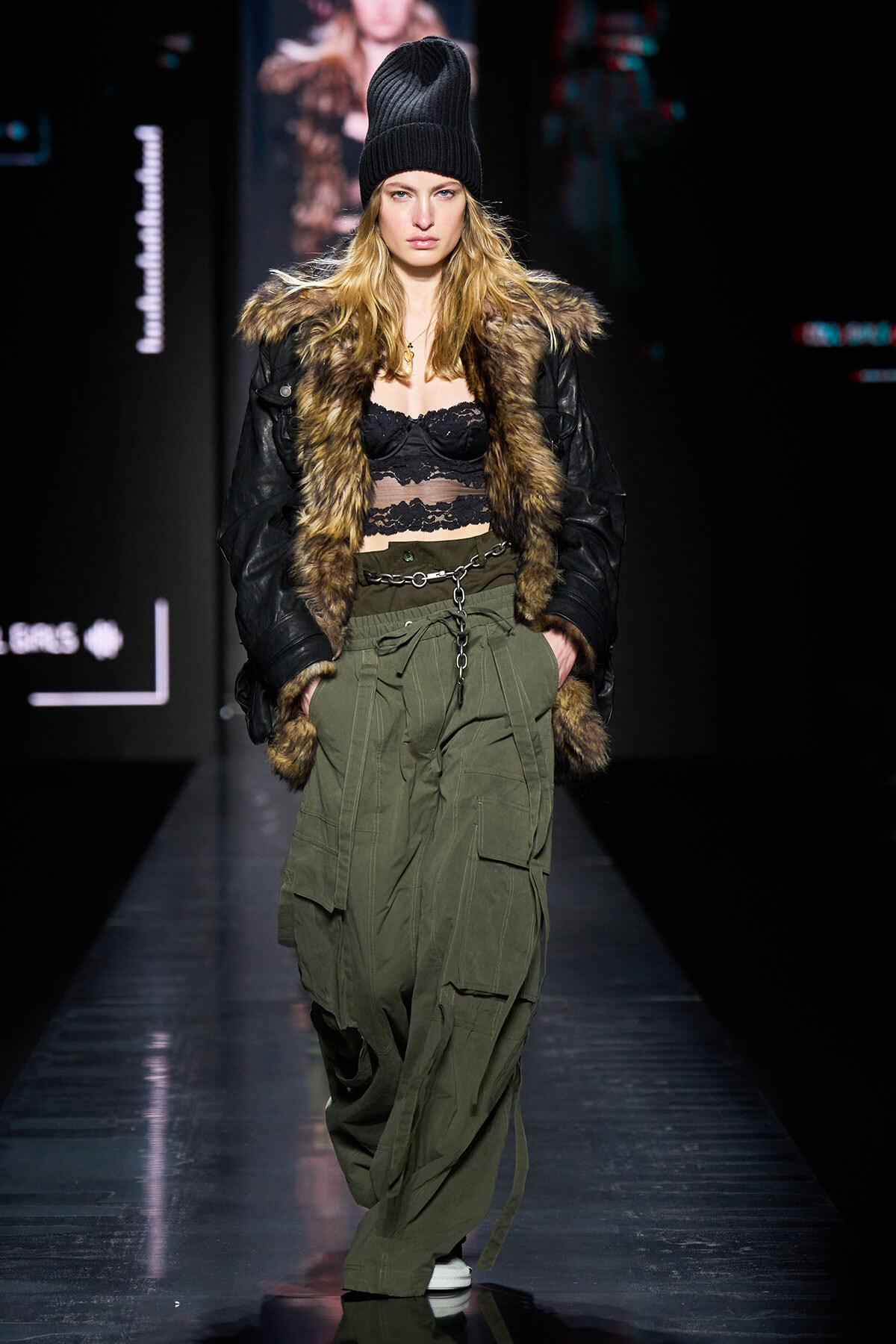 Model walking a fashion show in a black fur-trimmed jacket, lace crop top, and olive cargo pants with a metallic belt and beanie.
