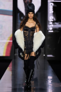 Female model walking the runway in a black corset-style dress with lattice cutouts, topped with a white faux-fur coat and a black beanie, paired with knee-high boots.