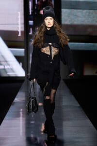 Model walking a fashion runway in an all-black ensemble: oversized knit cardigan, sheer lace top, beanie, and tall knit boots, carrying a black handbag.