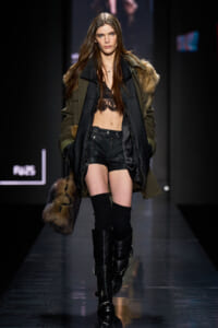 Model walking runway in an oversized olive parka with fur trim, black lace bralette, black leather shorts, knee-high boots, and a fur handbag edge.