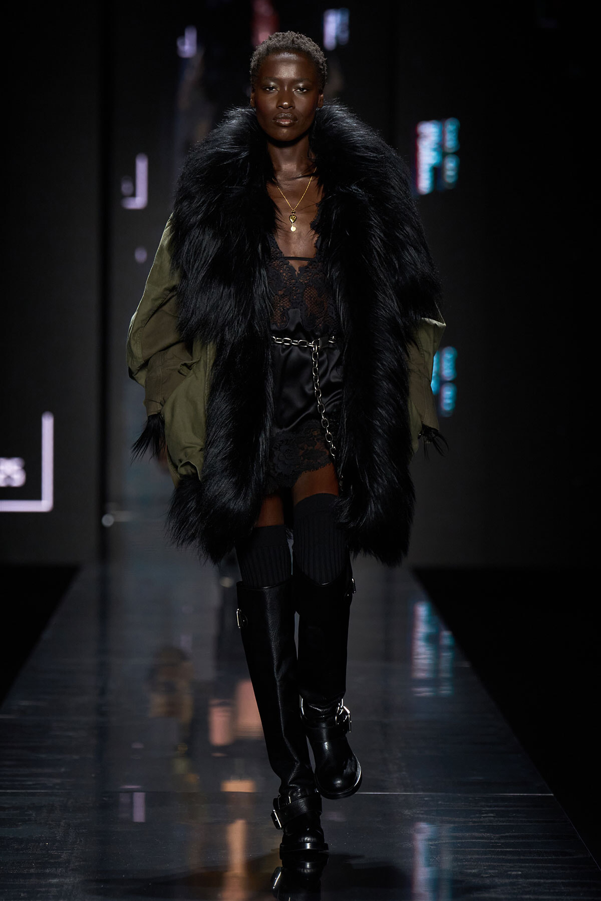 Model on a runway wearing a long black faux fur coat over a lacy black outfit with knee-high black boots and a chain belt.