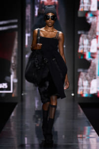 Model walks the runway in an all-black ensemble: strapless top, fur skirt, ankle boots, and a large black fur bag, with oversized sunglasses and a knit beanie.