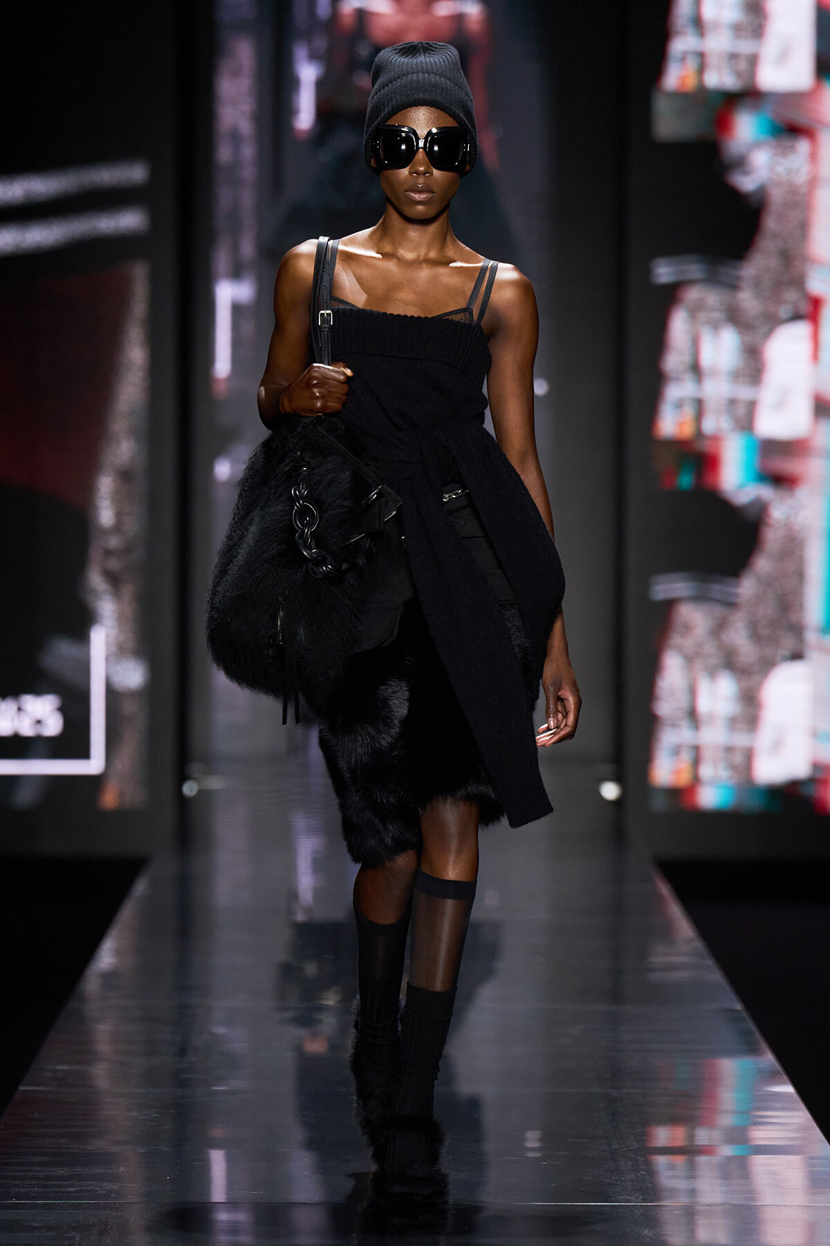 Model walks the runway in an all-black ensemble: strapless top, fur skirt, ankle boots, and a large black fur bag, with oversized sunglasses and a knit beanie.