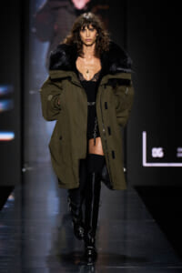 Model walks a fashion runway in an oversized olive parka with black fur collar, black lace top, and tall black buckled boots