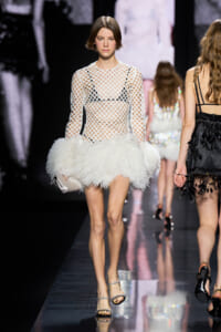 Model walking a fashion runway in a white fishnet long-sleeve top and fluffy white feathered mini skirt, holding a white clutch with silver sandals.