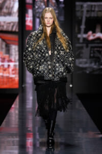 Model walking a runway in an oversized black quilted jacket adorned with silver beads and a long fringe skirt.