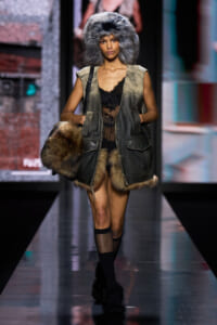 Runway model in a large gray fur hat, olive fur‑trimmed sleeveless vest, and sheer black lace top.