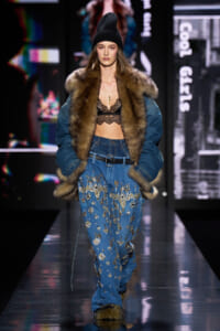 Model walks the runway in a fur-trimmed denim coat, lace bralette, and embellished denim jeans with a black beanie.