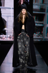 Model walks the runway in a long black coat with fur trim, distressed studded jeans, and a black beanie.
