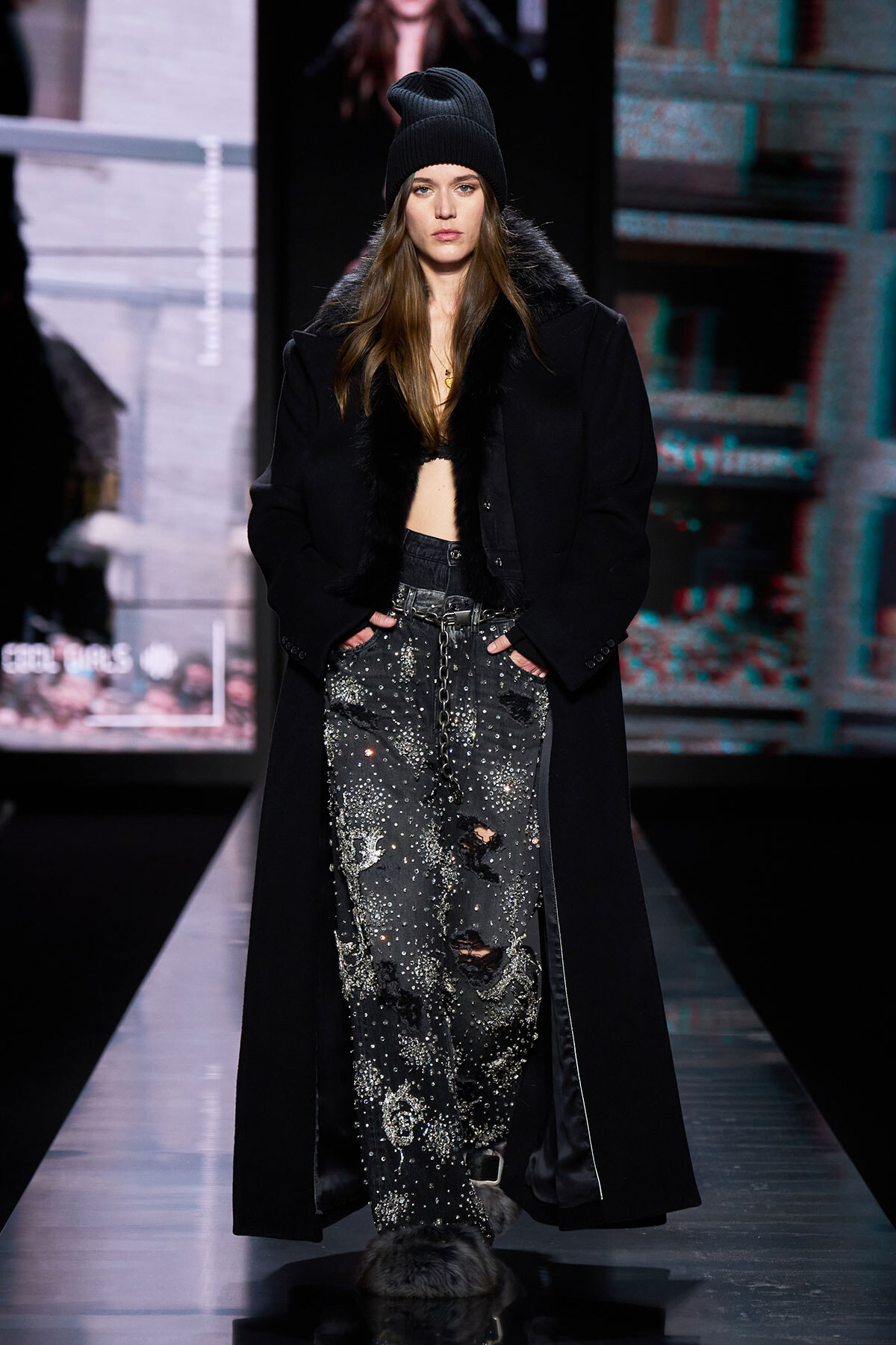 Model walks the runway in a long black coat with fur trim, distressed studded jeans, and a black beanie.