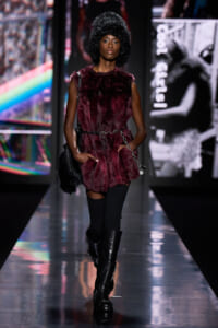 Model on a glossy runway wearing a burgundy faux-fur vest, black thigh-high socks, and tall black boots with a chain belt and oversized afro hat.