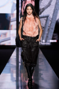 Model walking a fashion runway in a peach fur sleeveless top, dark cargo pants, black boots, and a black shoulder bag.