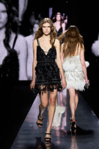 Model walks down a glossy runway wearing a black fringed dress and strappy heels at a fashion show.