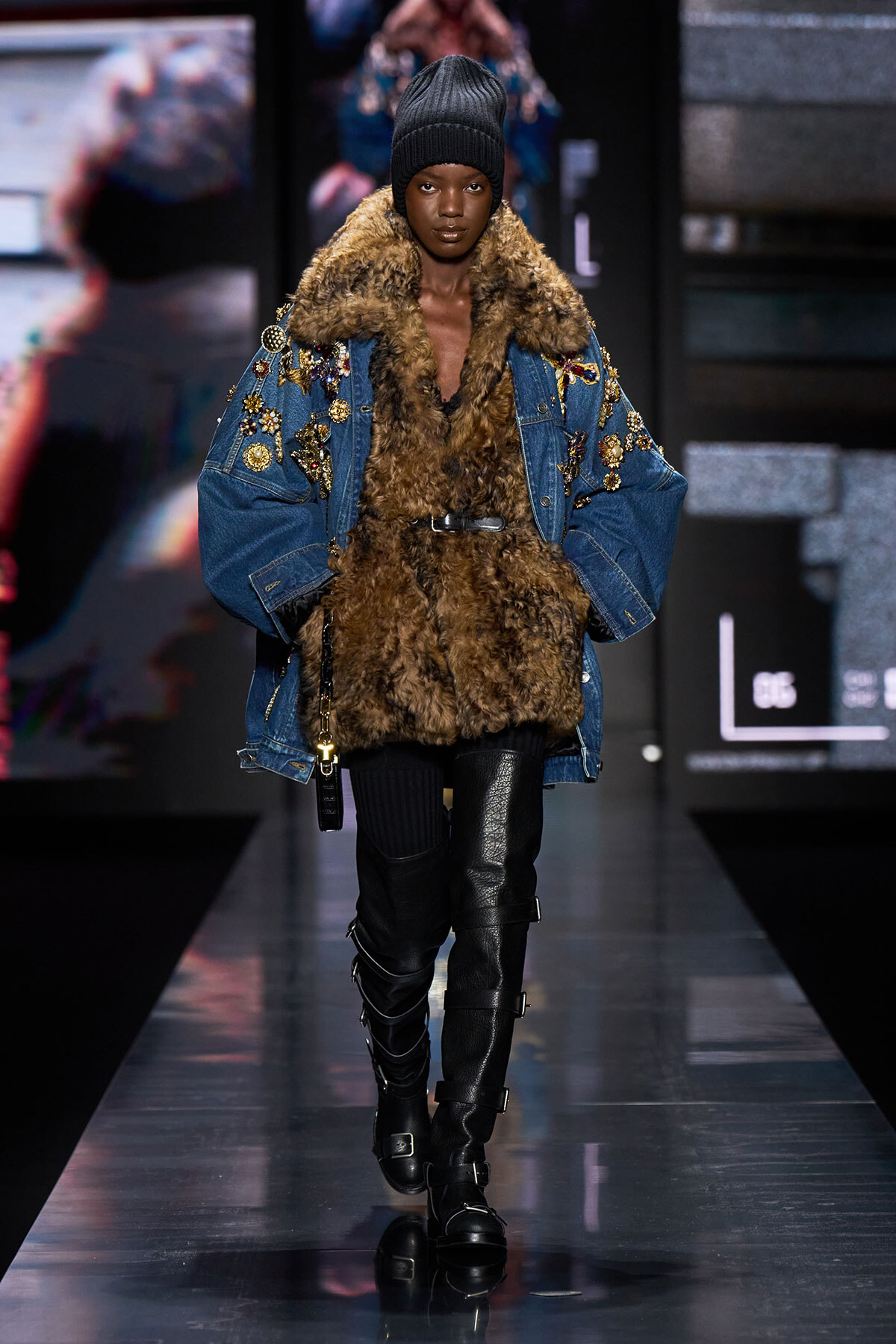Runway model wears a fur-trimmed brown coat under a decorated denim jacket with floral embroidery, paired with a black knit beanie and tall buckled boots.