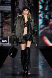Model walking the runway in an oversized olive bomber, black lace bralette and shorts, and tall buckled boots with a black knit beanie.
