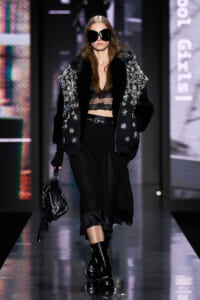 Model walking the runway in a black embellished fur-trim jacket, sheer lace top, and black skirt, carrying a black handbag and wearing oversized sunglasses.