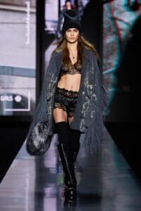 Model on a runway wearing a black knit beanie, a gray fringe coat, black lace lingerie with matching shorts, black thigh-high socks and boots, and a gray fur bag.