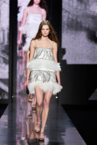 Model walks a runway in a white fringe mini dress with silver beading and feather trim, carrying a small black clutch.