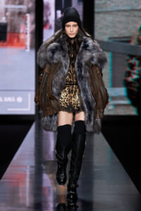 Model walks the runway in a oversized brown fur coat with a gray fur collar, leopard print dress, black beanie, and tall black boots on a glossy catwalk.